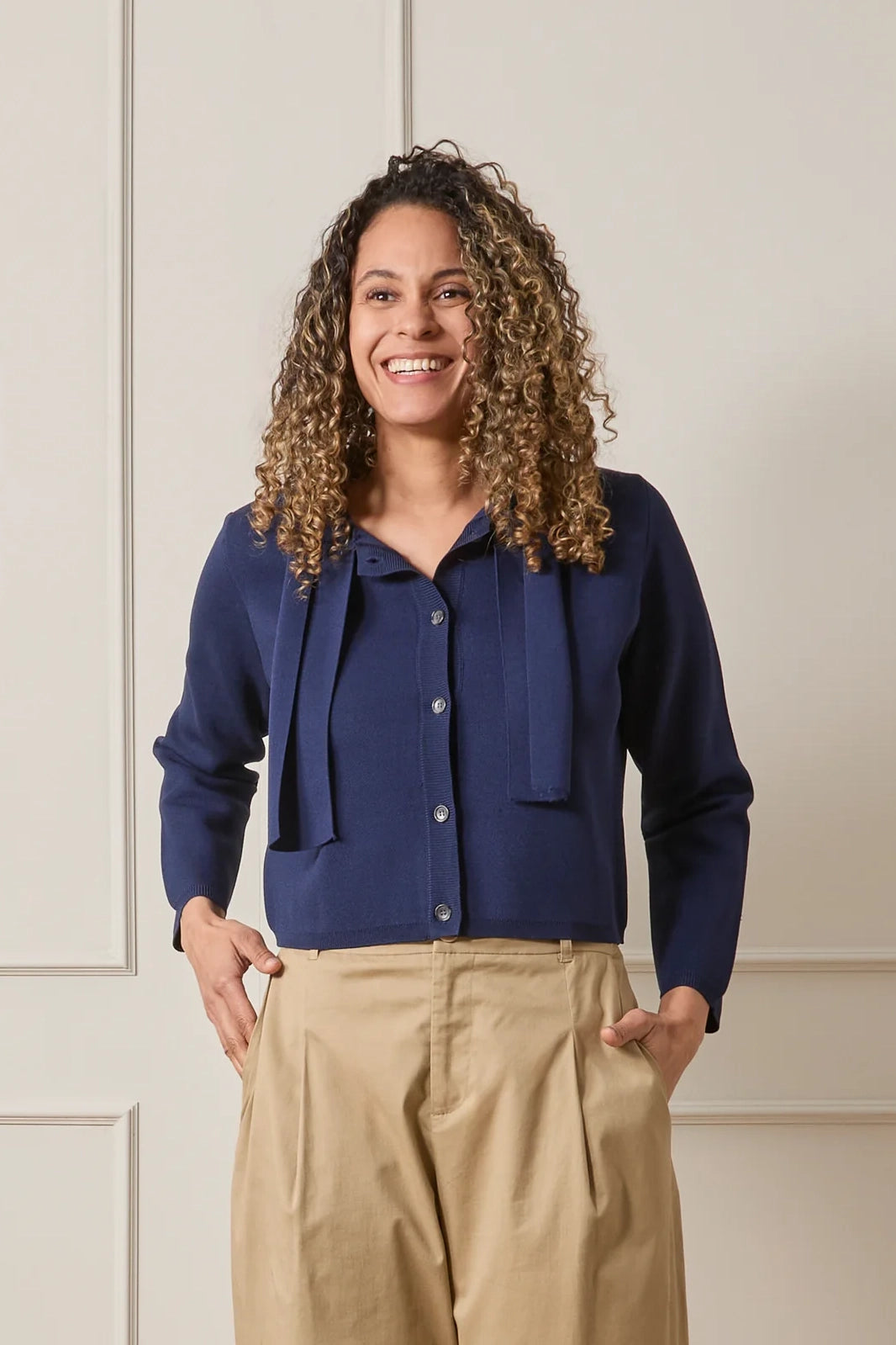 Woman wearing a blue blouse with a bow and beige pants against a neutral background