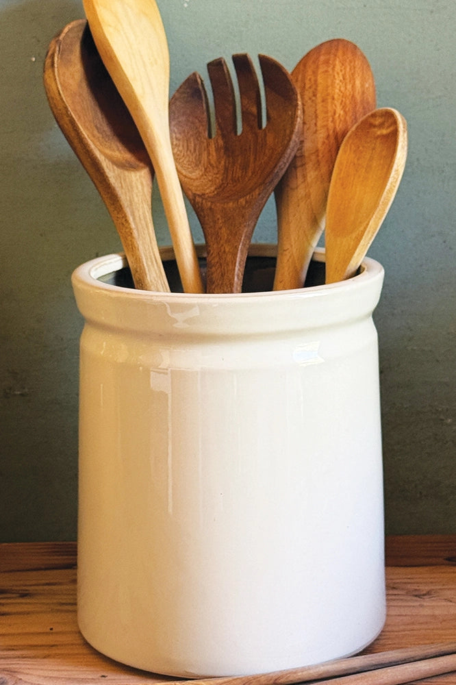 A white ceramic crock pot container holding several wooden kitchen utensils, including spoons and a spatula, set on a wooden surface against a dark green wall.