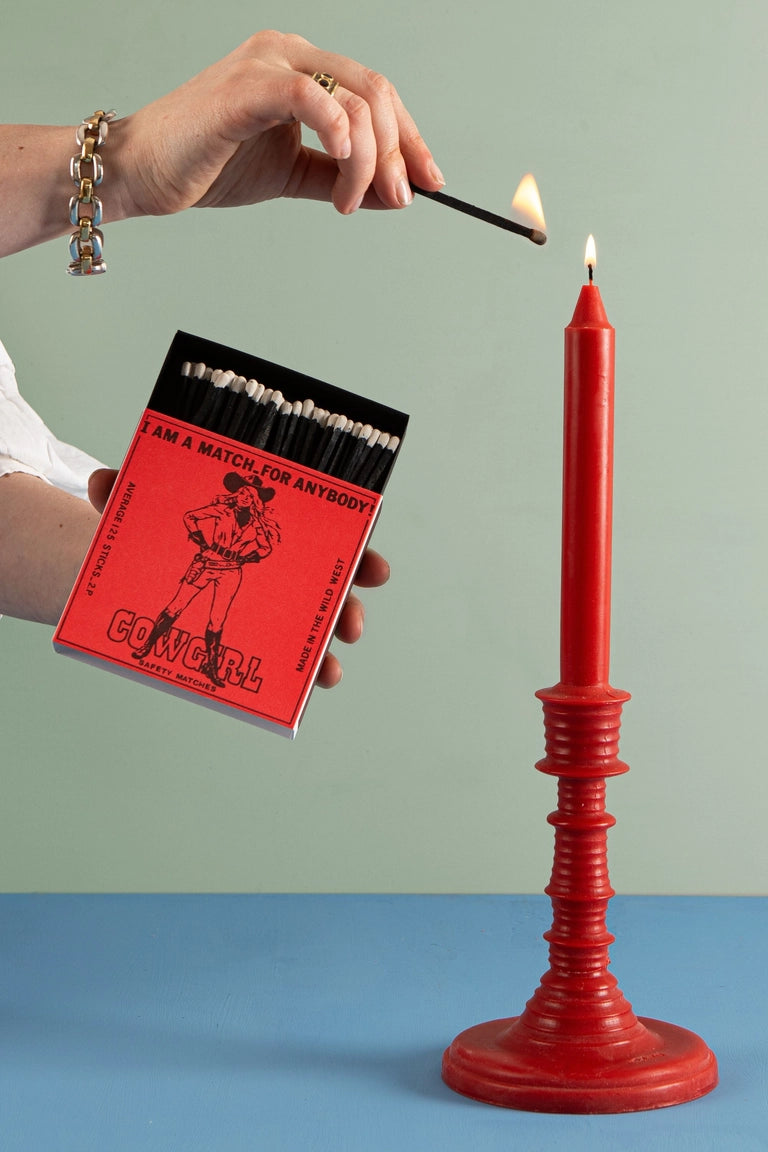 Person lighting a red candle with a matchbook labeled 'Cowgirl' against a green and blue gradient background.