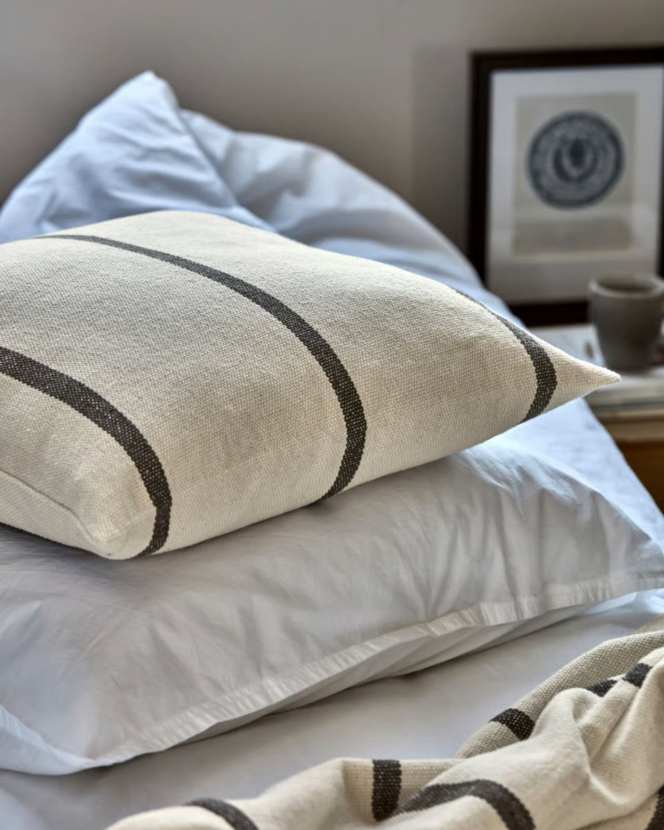 A close-up of a textured linen throw pillow in a warm off-white color with dark charcoal stripes, resting on white bedding in a softly lit bedroom setting.