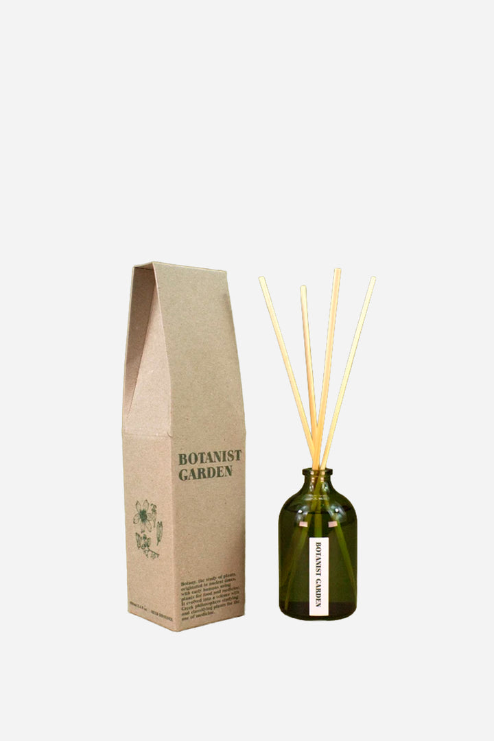 A reed diffuser set with an olive green glass bottle, natural wood reeds, and a brown kraft paper box labeled 'BOTANIST GARDEN' on a light gray background.