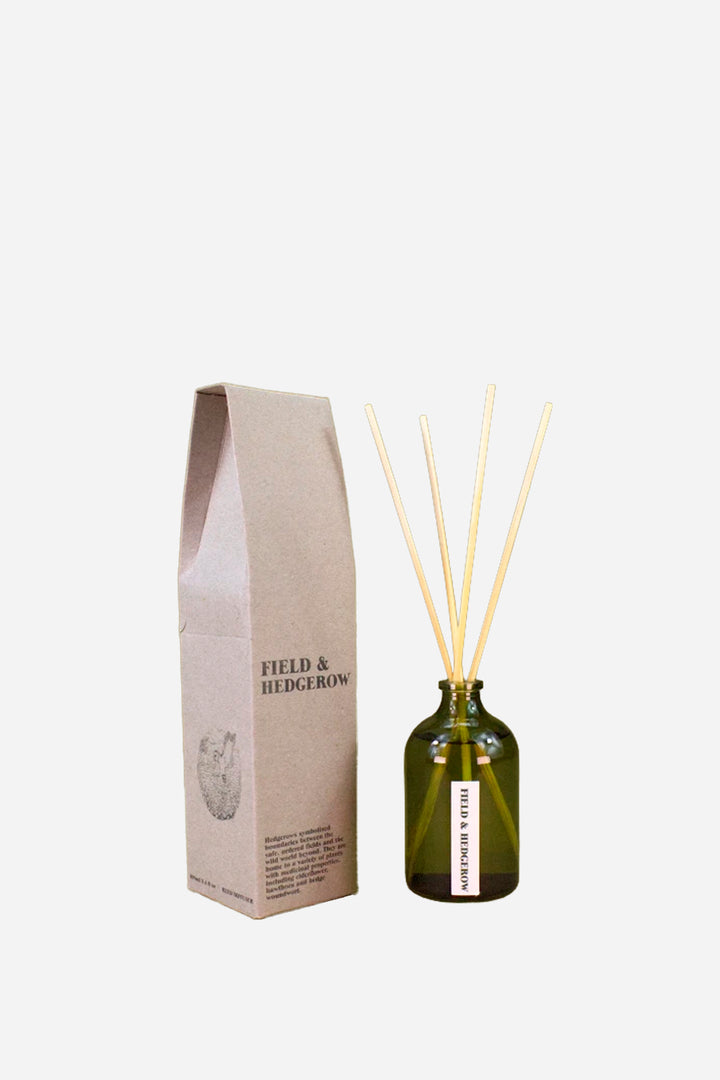 A reed diffuser bottle made of dark olive green glass with natural light wood reeds, standing next to its kraft paper packaging box. The box is printed with the brand name 'FIELD & HEDGEROW'.