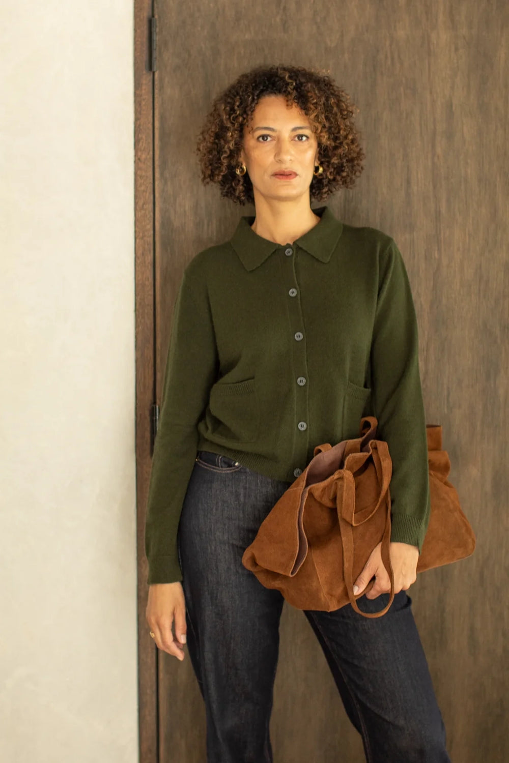 Woman wearing a green cardigan and holding a brown leather bag against a wooden wall.