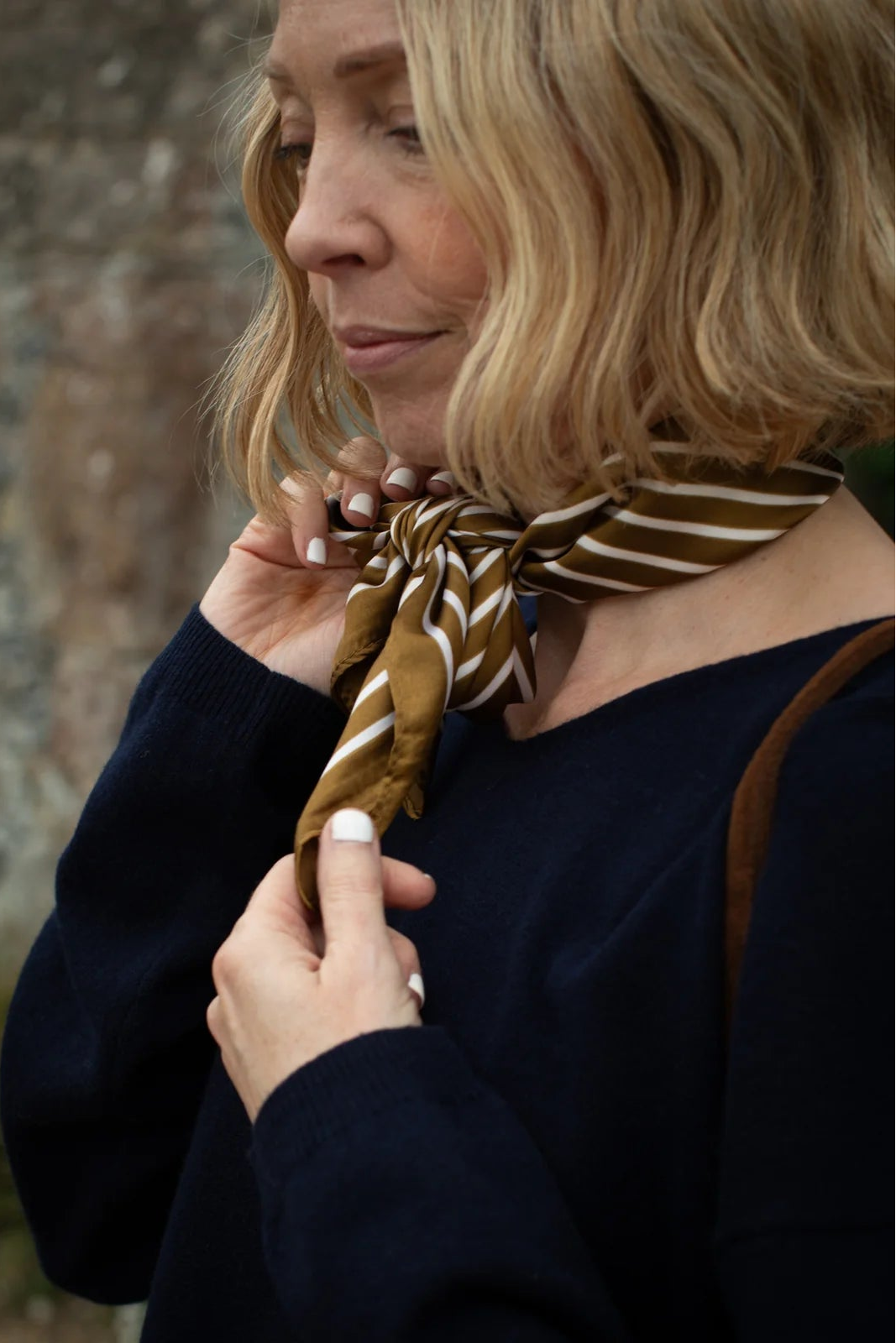 Woman adjusting a brown and gold striped scarf against a stone wall background
