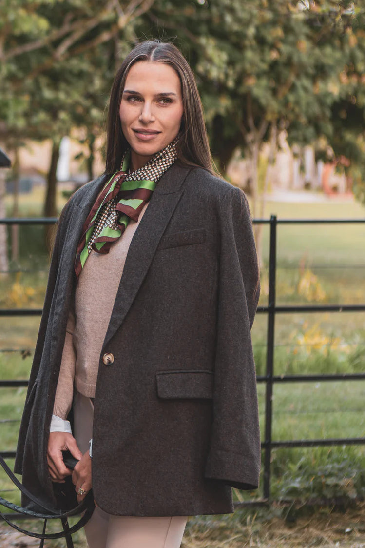 Woman wearing a brown blazer and scarf outdoors with a fence and trees in the background