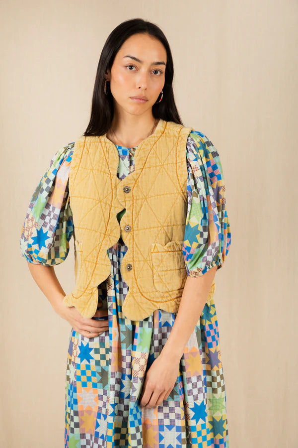 Woman wearing a yellow quilted vest over a colorful patterned dress on a beige background