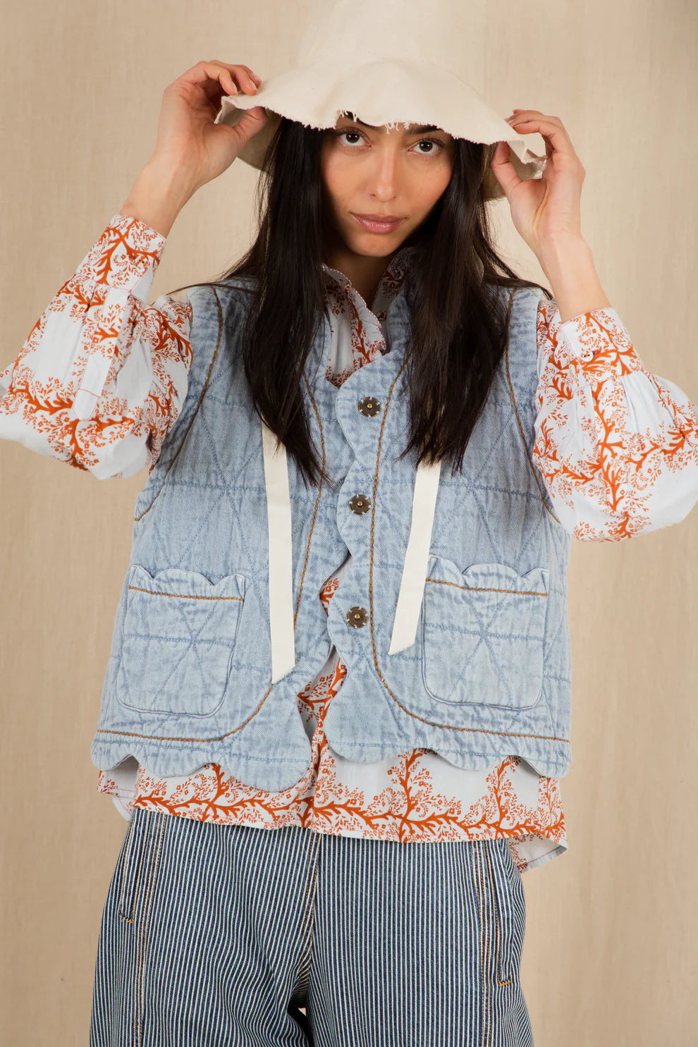 Woman wearing a denim vest with floral lace trim and a white hat against a beige background