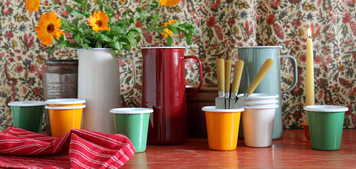 collection of colourful falcon enamelware set against floral backdrop with vintage cutlery and vase of flowers