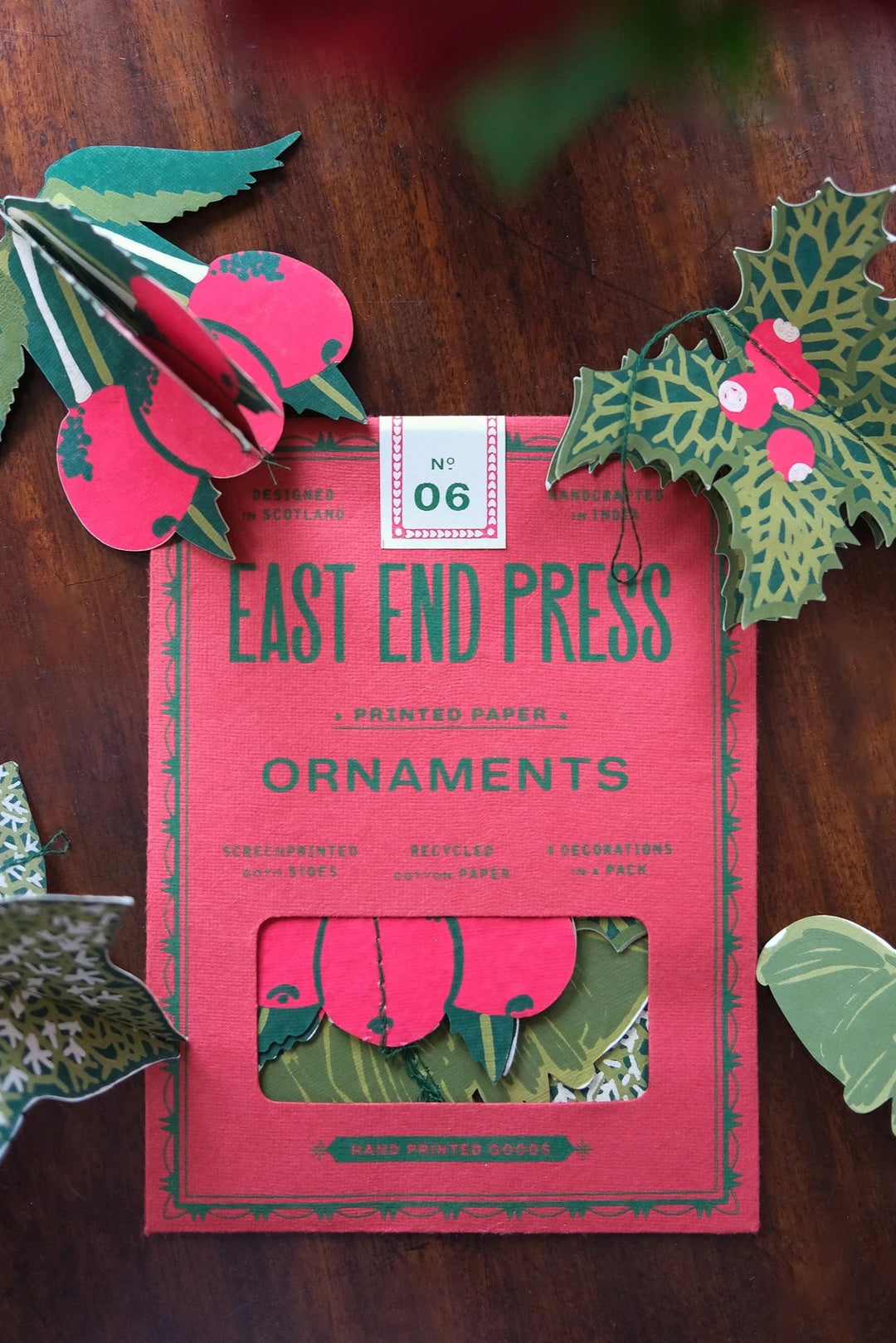 Pack of East End Press ornaments with decorative paper holly leaves and berries on a wooden surface.