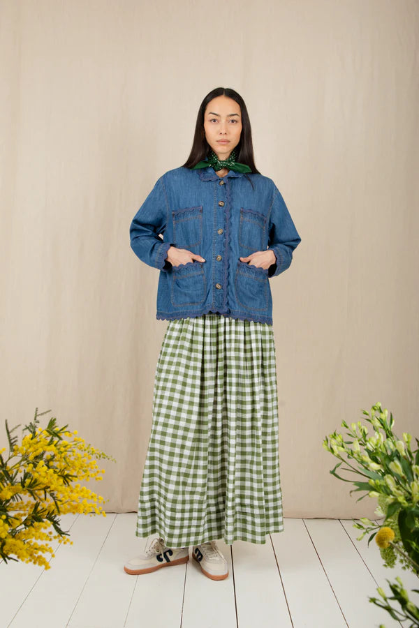 Woman wearing a blue denim jacket and green checkered dress standing against a beige wall with floral decorations.