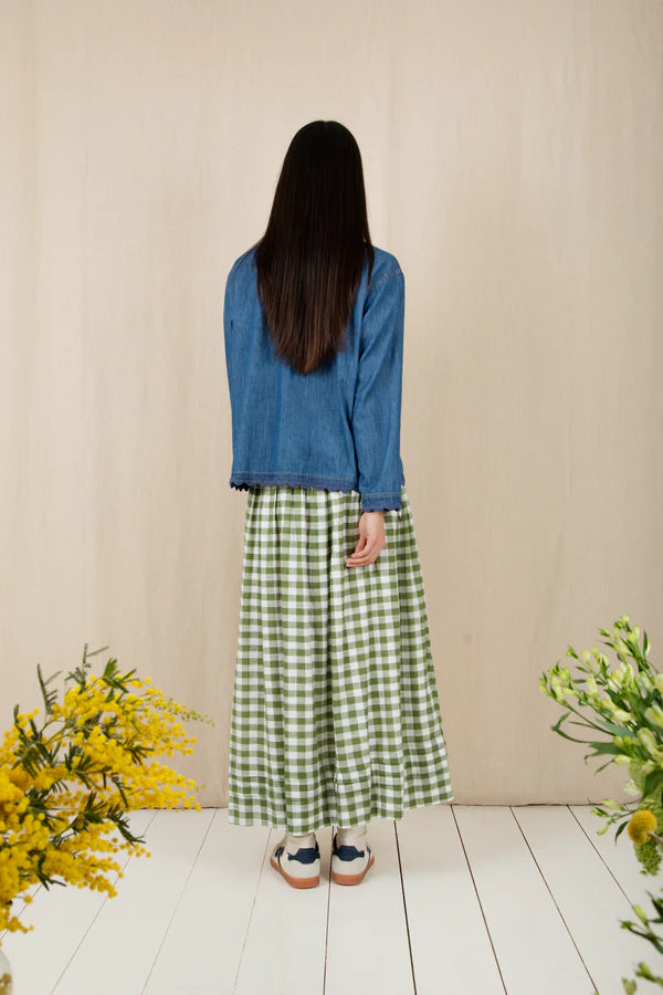 Person wearing a blue top and green checkered skirt standing in front of a beige wall with flowers on either side.