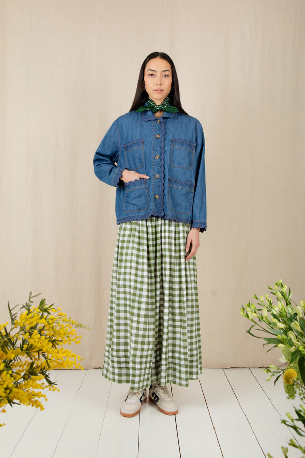 Woman wearing a blue denim jacket and green checkered pants standing against a beige wall with floral decorations.
