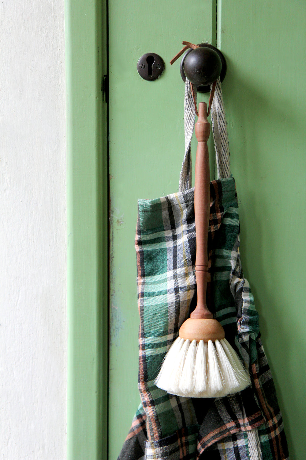 lifestyle image of Redecker Goat Hair Duster with wooden handle, hanging on green utility door with checked apron