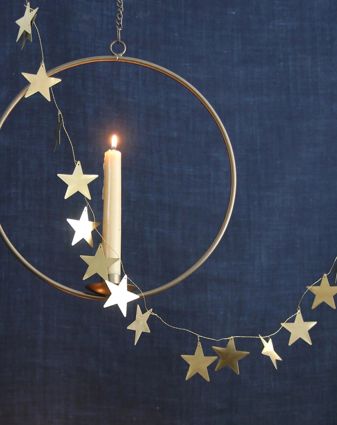 metal wire garland strung with brass Christmas stars pictured with a hanging candle ring and lit beeswax candle