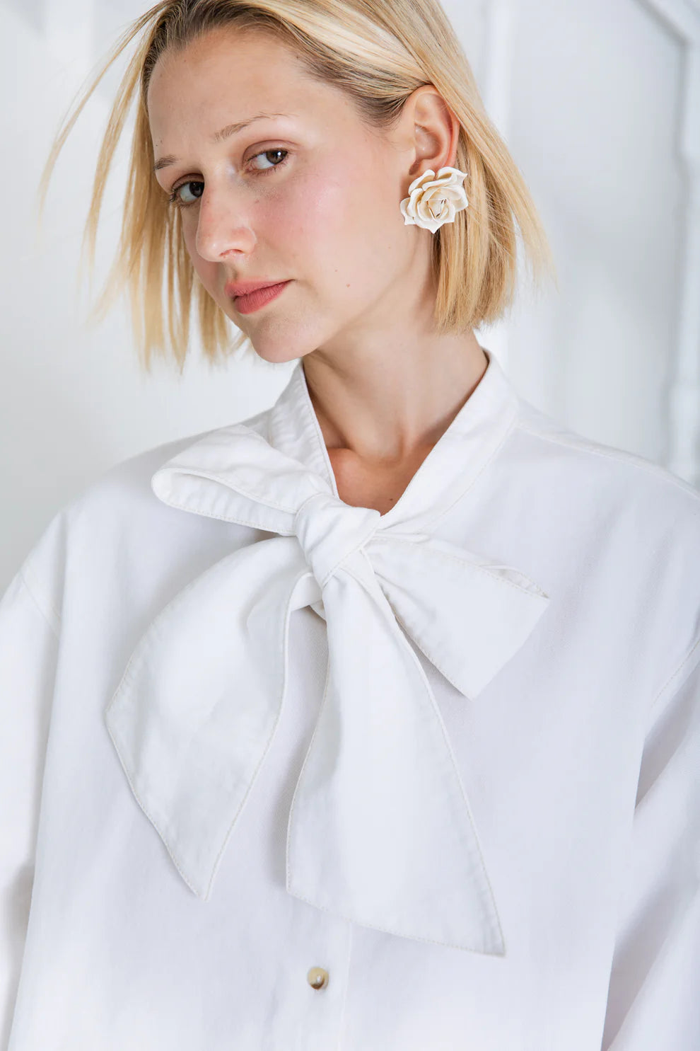 Woman wearing a white coat with a large bow and floral earrings against a light background