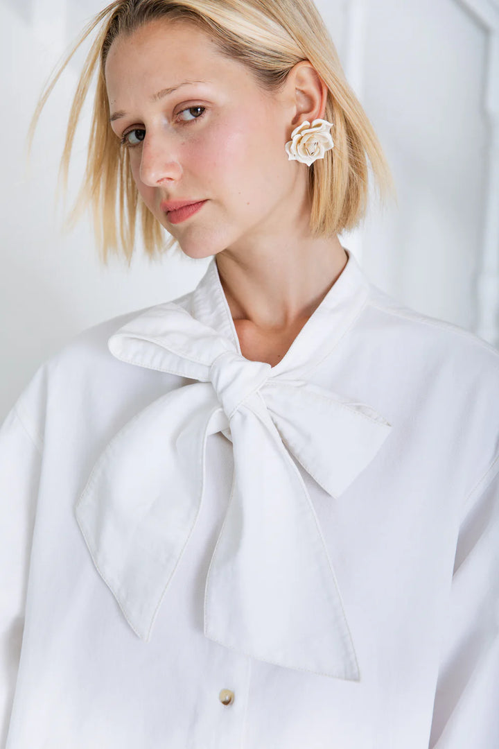 Woman wearing a white coat with a large bow and floral earrings against a light background