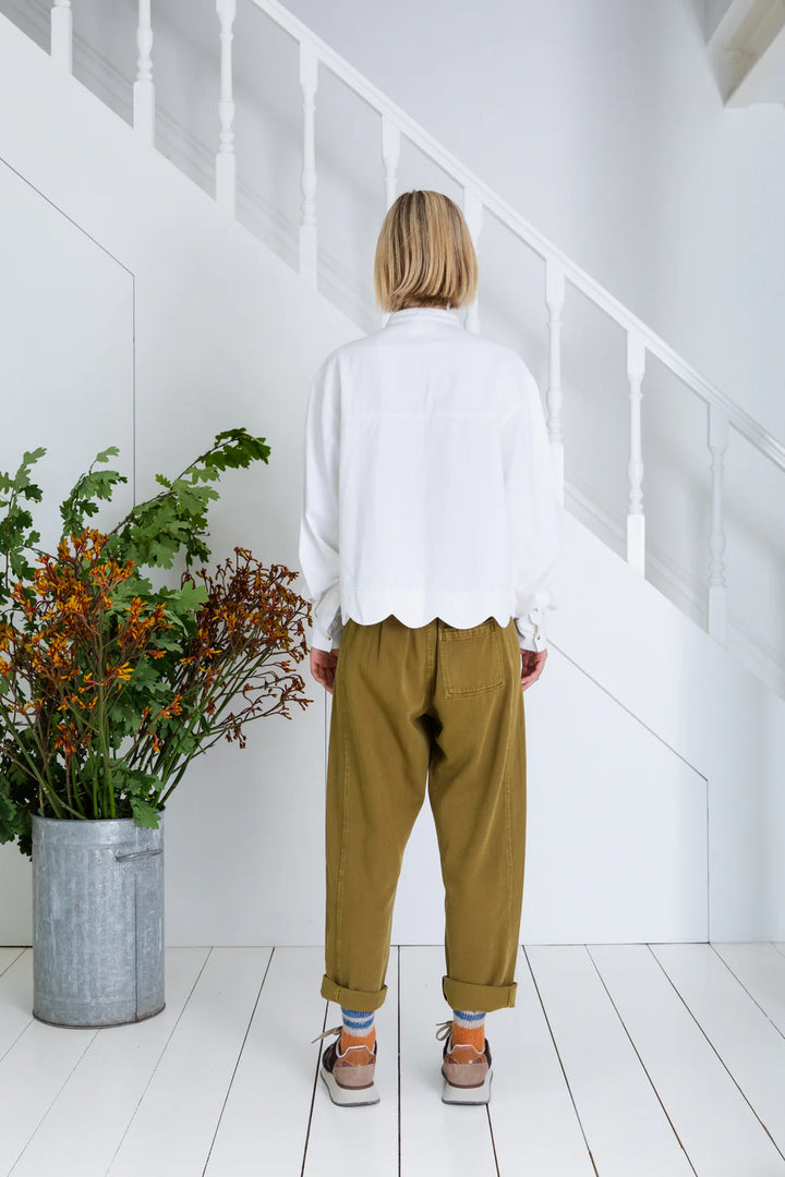 Person wearing a white shirt and mustard pants standing in a minimalistic room with a plant.