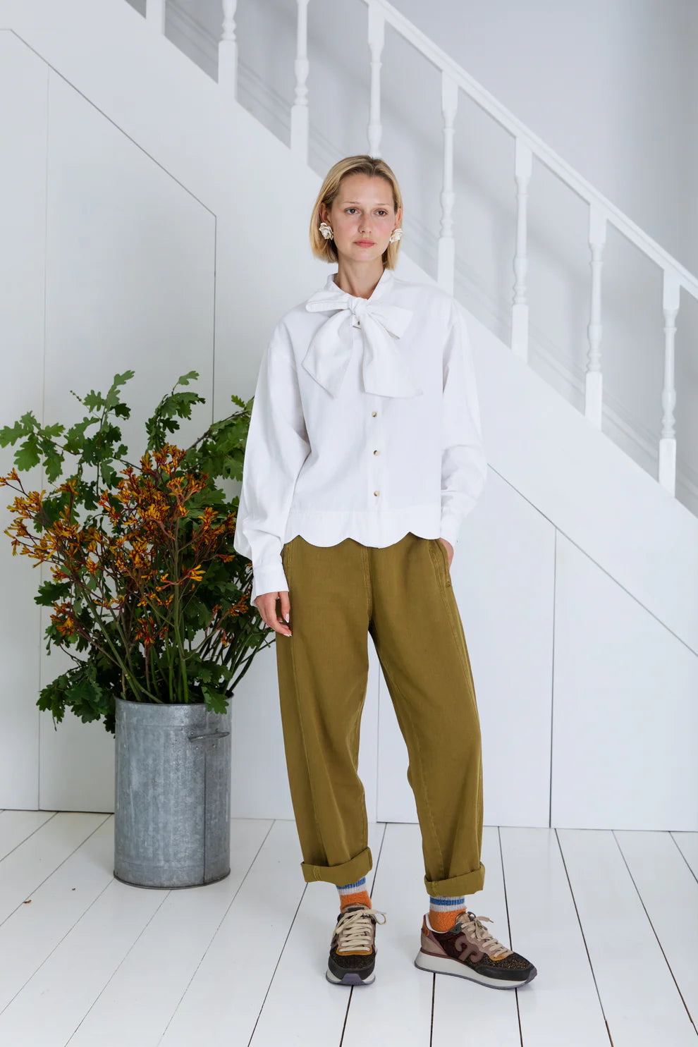 Person wearing a white blouse with a large bow and green pants standing in a minimalistic room with a plant.