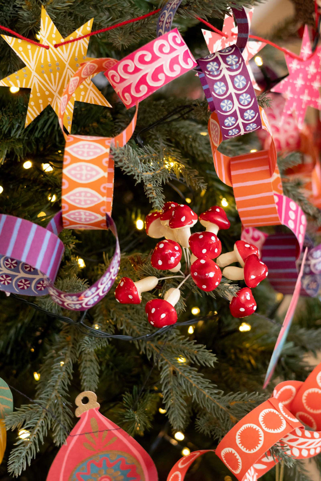 A close-up of a decorated Christmas tree with colorful patterned paper ribbons, red and white mushroom ornaments, and warm white string lights.