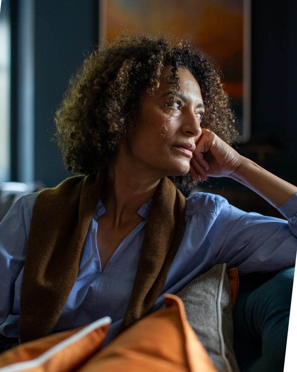 Woman sitting on a couch with an orange pillow, looking thoughtful.