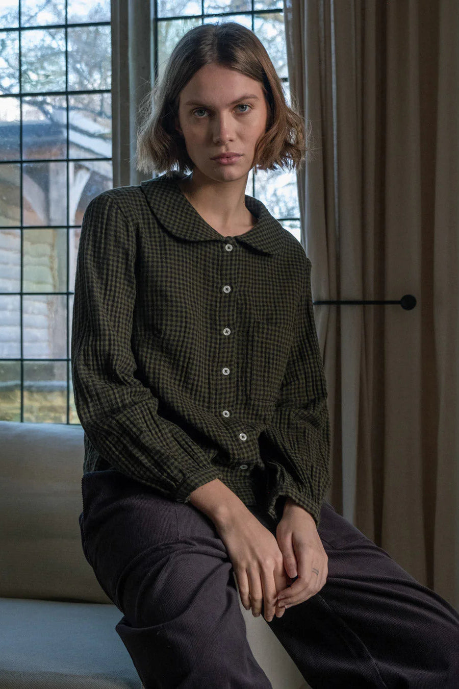 Woman sitting indoors wearing a green checkered blouse and dark pants.