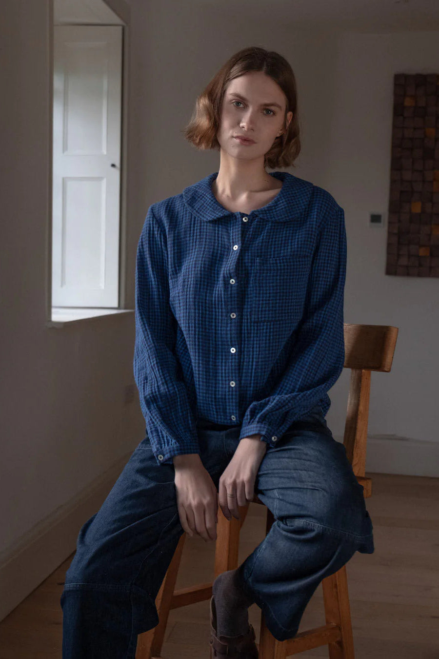 Person wearing a blue checkered shirt and pants sitting on a wooden chair in a room with a window and wall art.