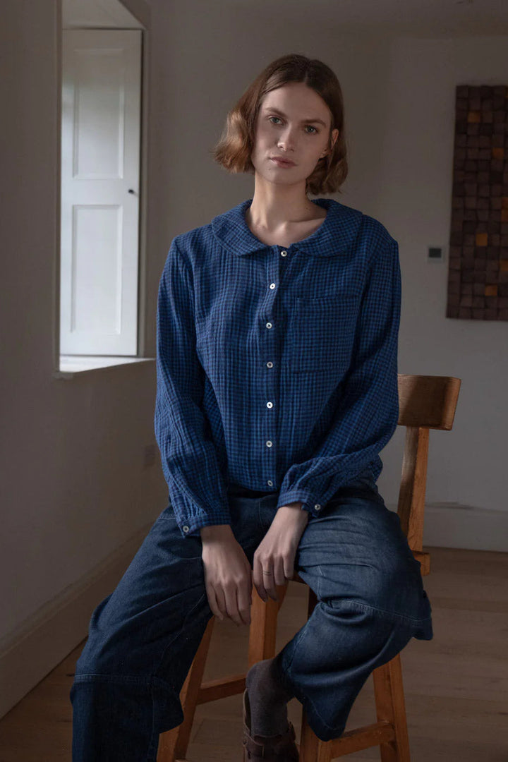 Person wearing a blue checkered shirt and pants sitting on a wooden chair in a room with a window and wall art.