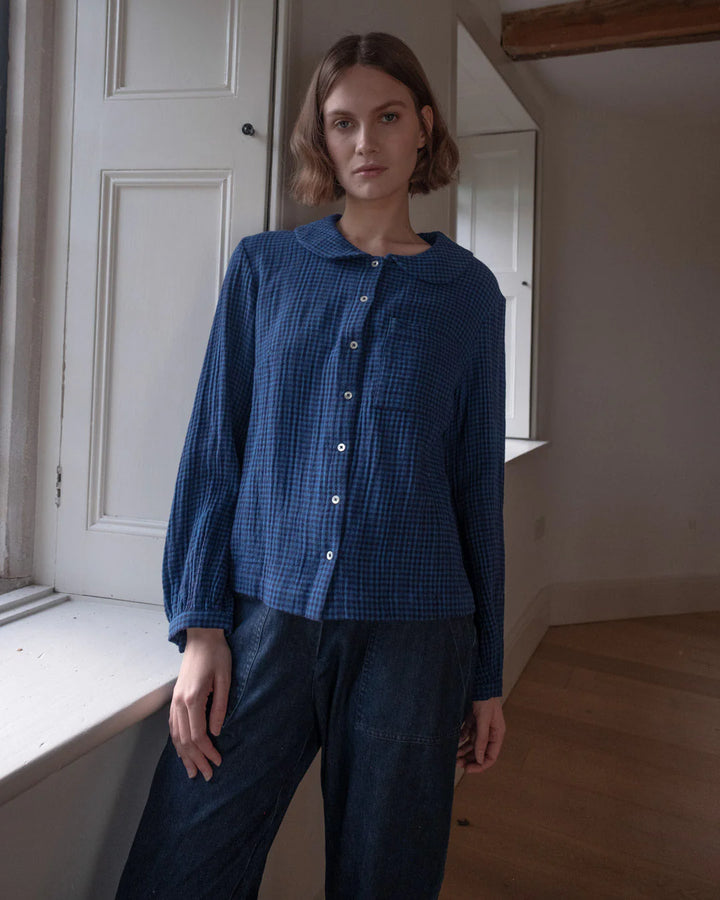 Woman wearing a blue checkered shirt and dark pants standing in a room with white walls and wooden floor.