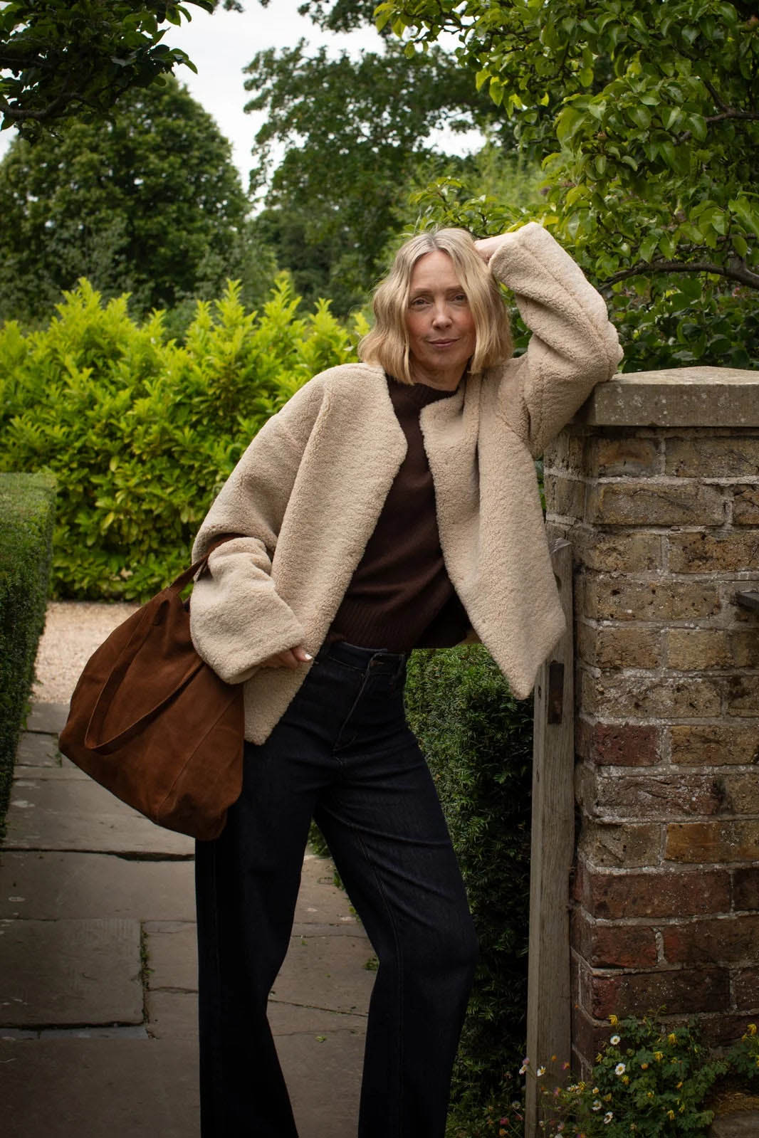 Woman in a beige coat and brown bag standing against a brick wall with greenery in the background