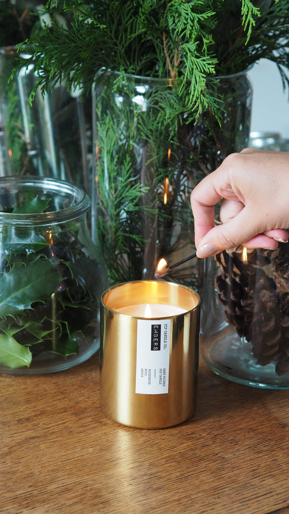 Gold candle holder with a label being held over a glass vase with greenery on a wooden surface.