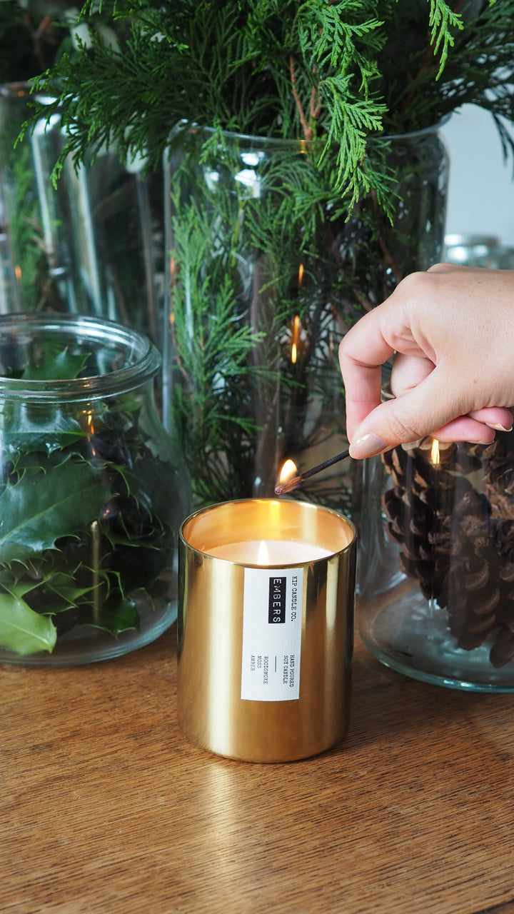 Gold candle holder with a label being held over a glass vase with greenery on a wooden surface.