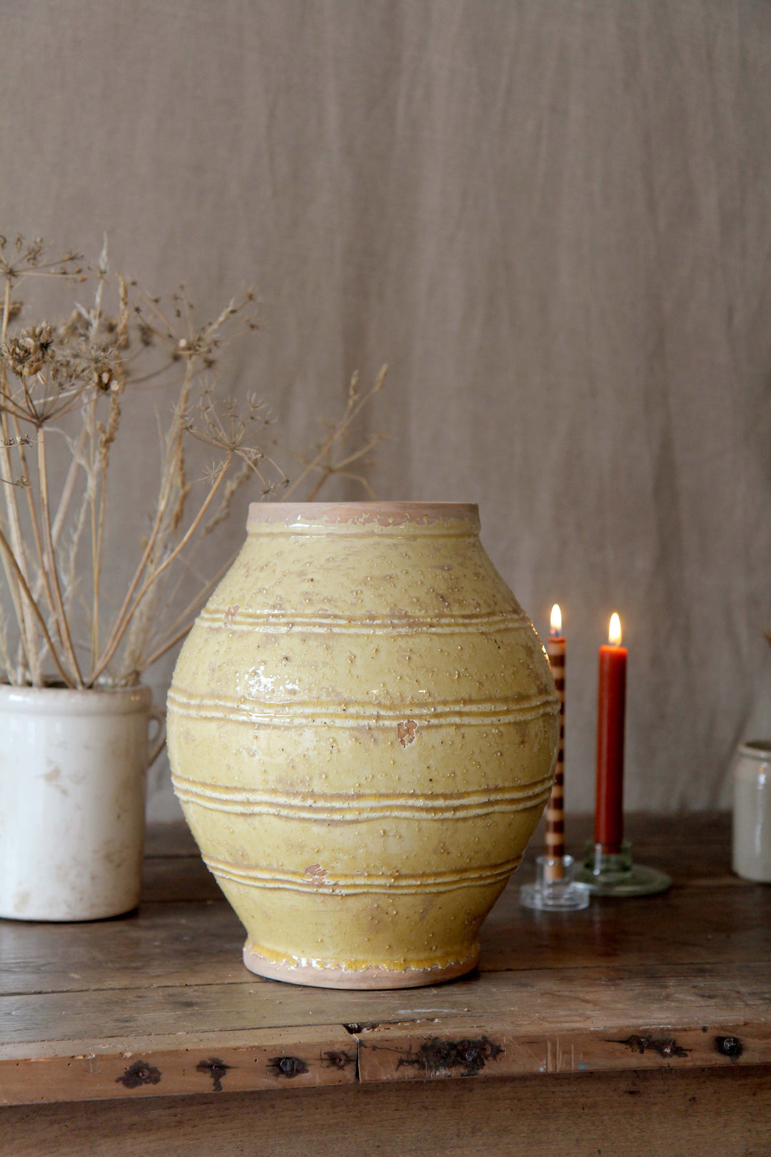 Yellow ceramic vase on a wooden surface with candles and plants in the background