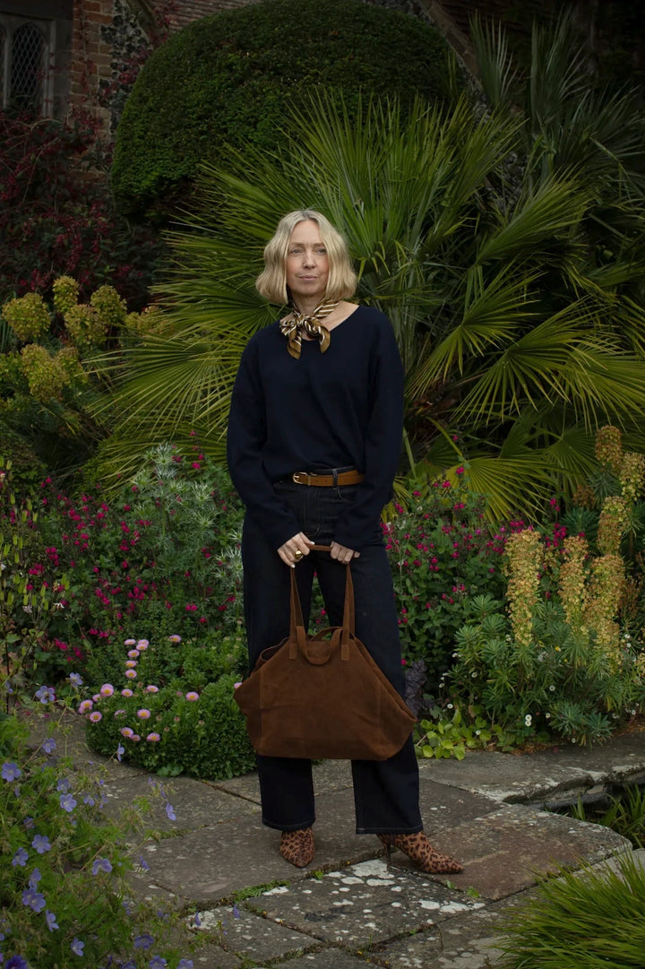 Woman standing in a garden holding a brown bag, surrounded by greenery and flowers.
