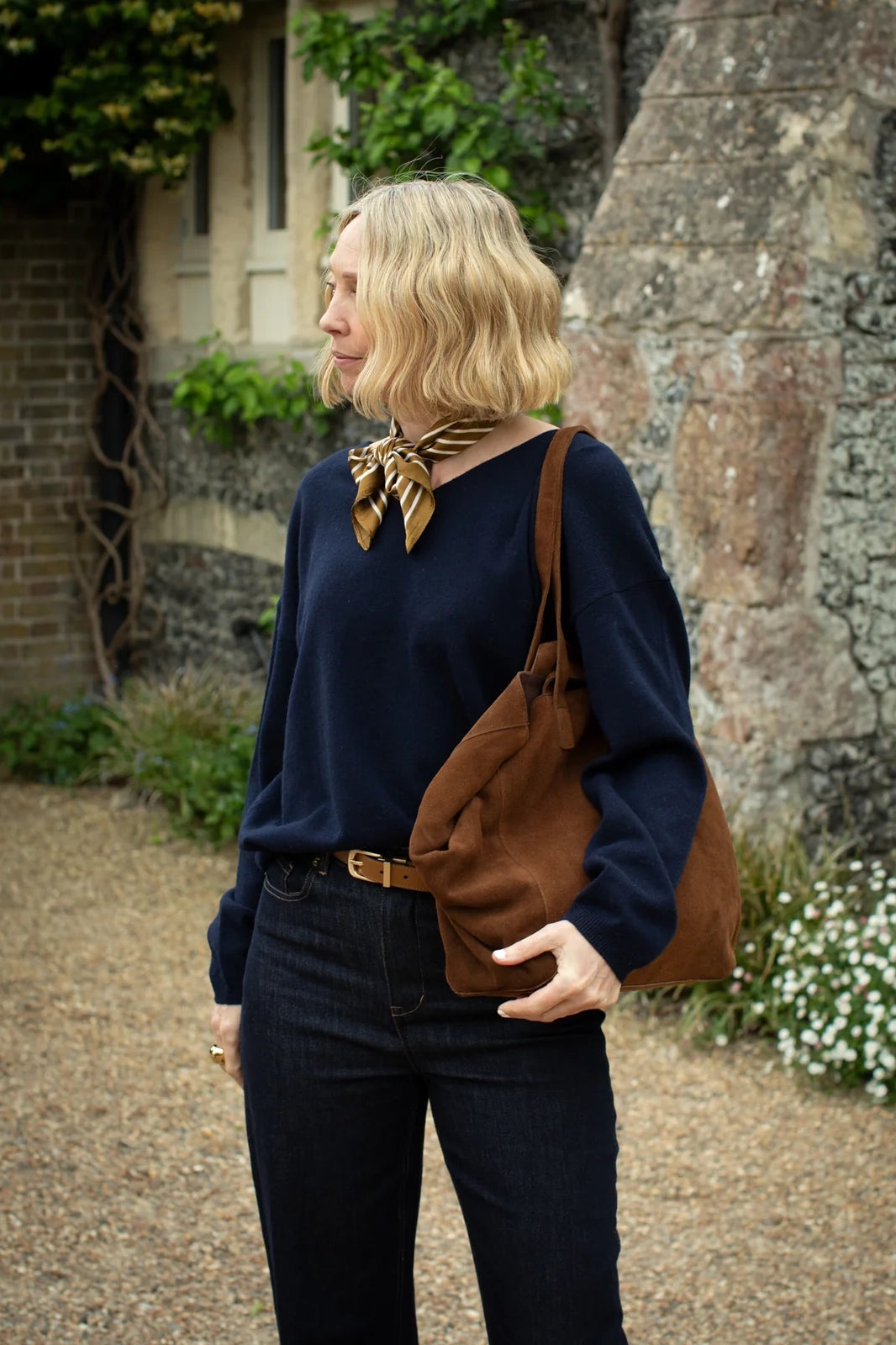 Woman holding a brown leather bag outdoors with a stone building and greenery in the background