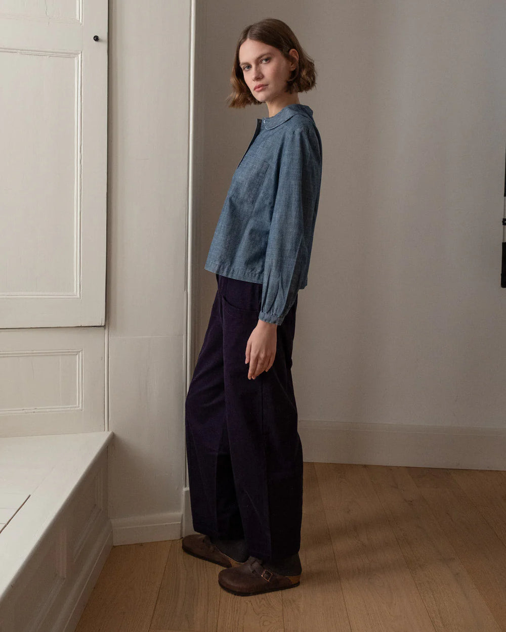 Woman wearing a blue denim shirt and navy cord trousers standing in a room with white walls and wooden floor.