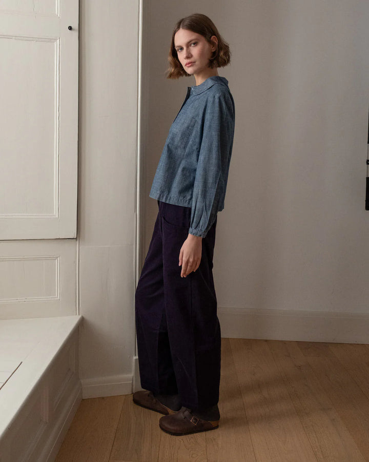 Woman wearing a blue denim shirt and navy cord trousers standing in a room with white walls and wooden floor.