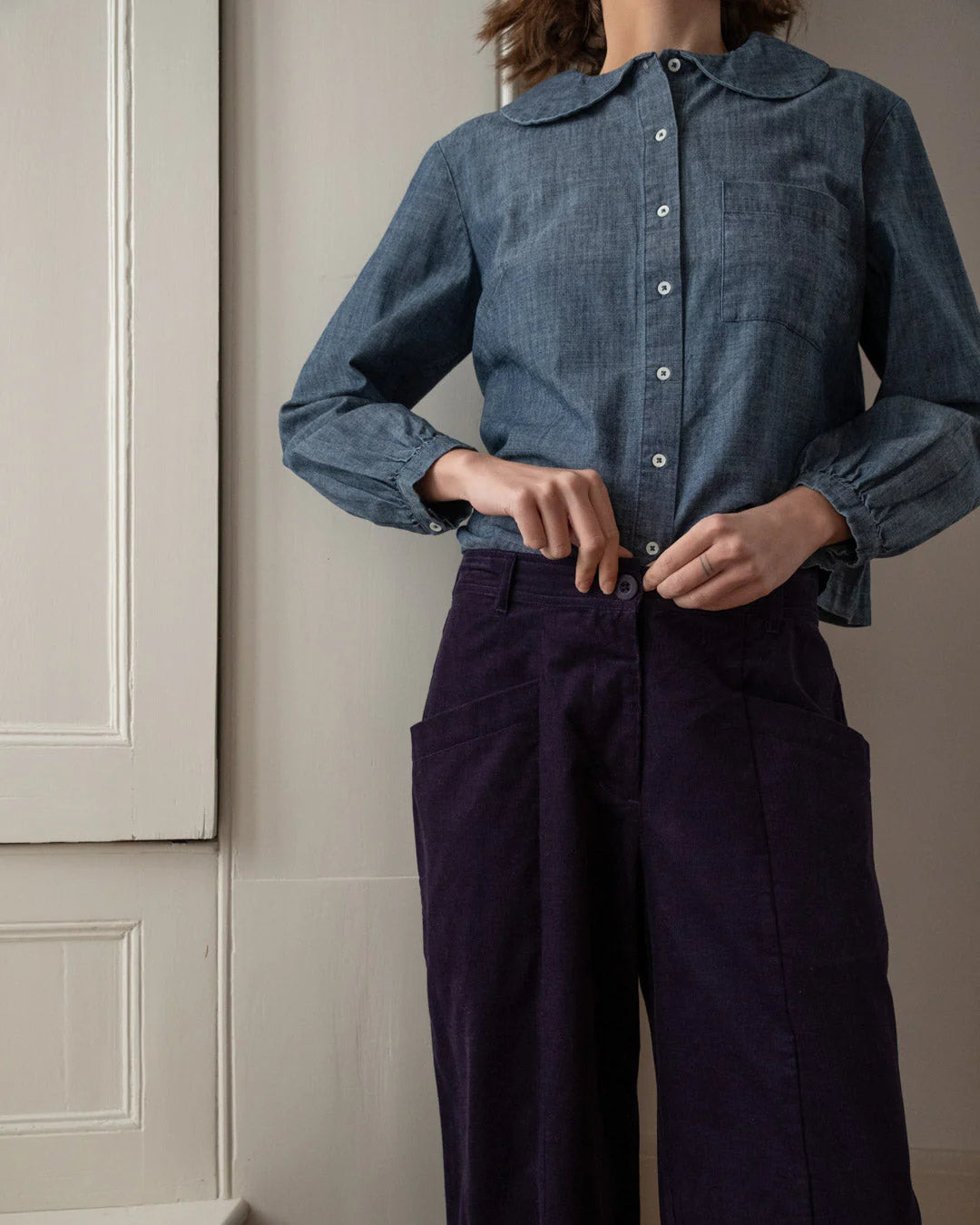 Person wearing a blue denim shirt and dark pants against a neutral wall.