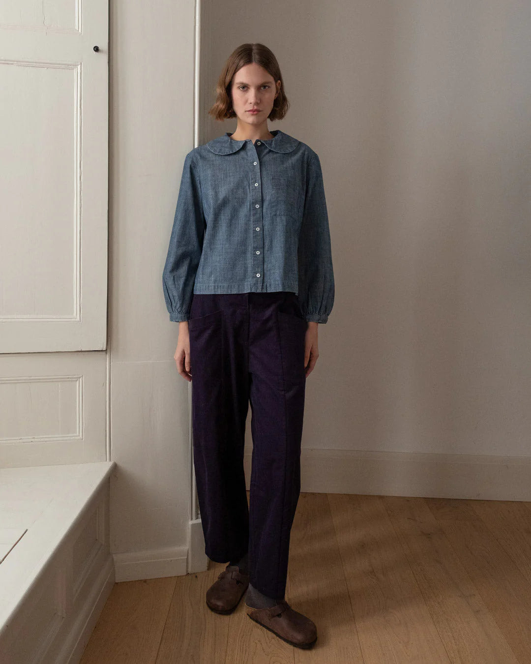 Person wearing a blue checkered shirt and dark pants standing in a room with a neutral wall and wooden floor.