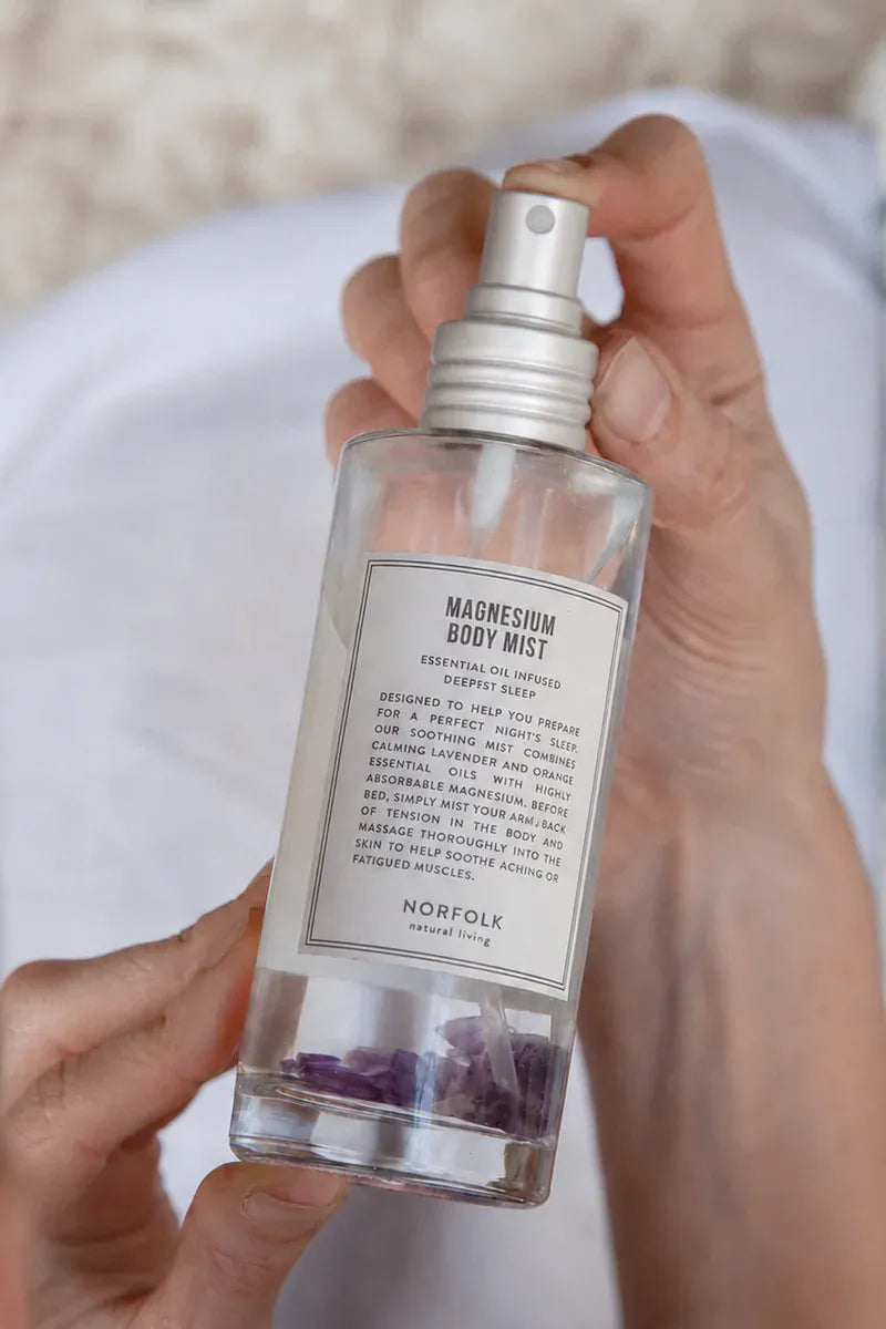 A hand holding a clear spray bottle of Norfolk Magnesium Body Mist with a white label and silver pump, set against a soft white background.