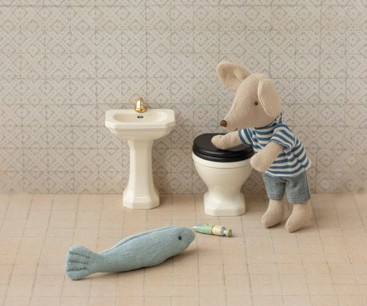Stuffed toy mouse on a toilet with a fish and roll of toilet paper on the floor, in front of a sink.