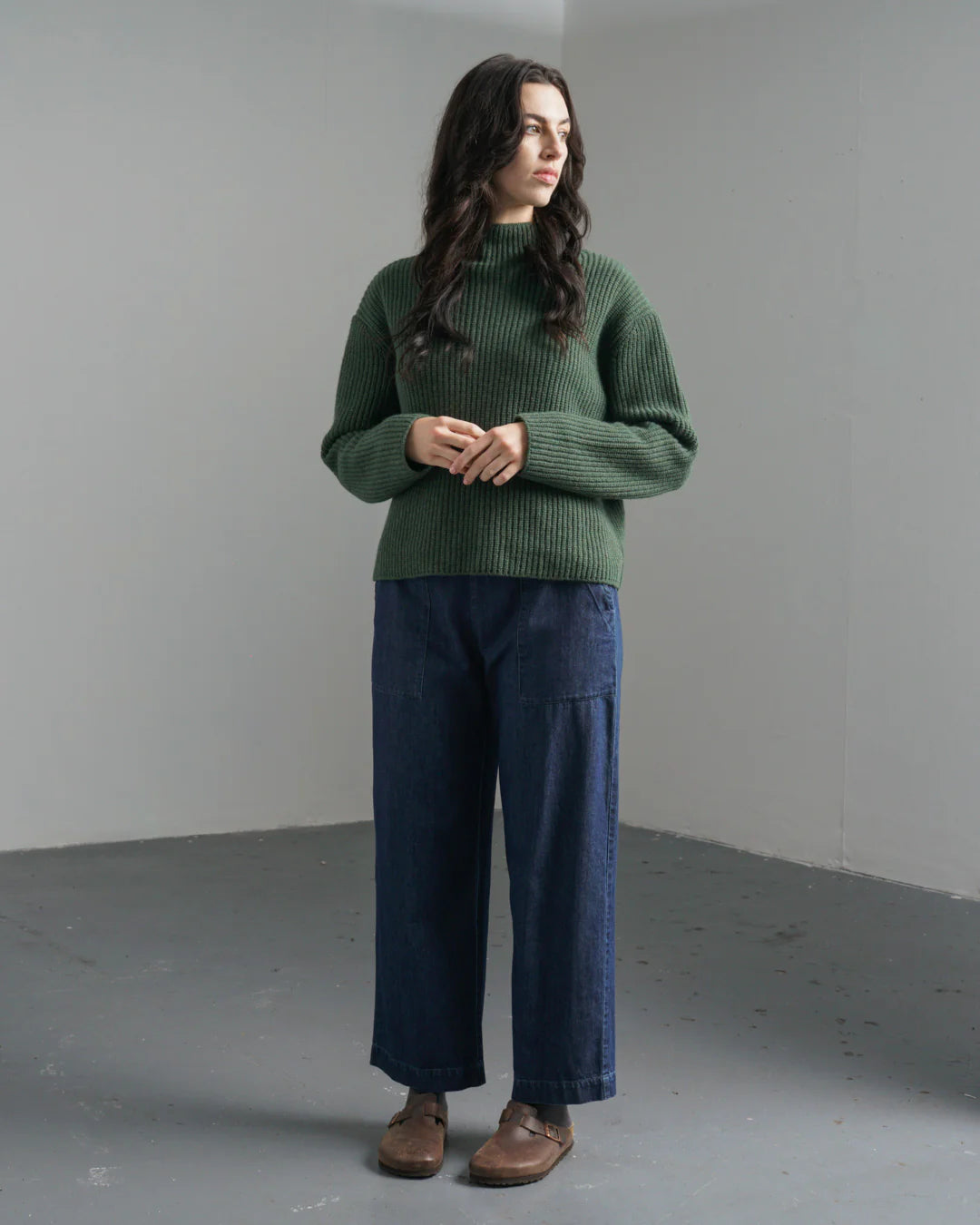 Woman wearing a green sweater and blue pants standing against a plain background