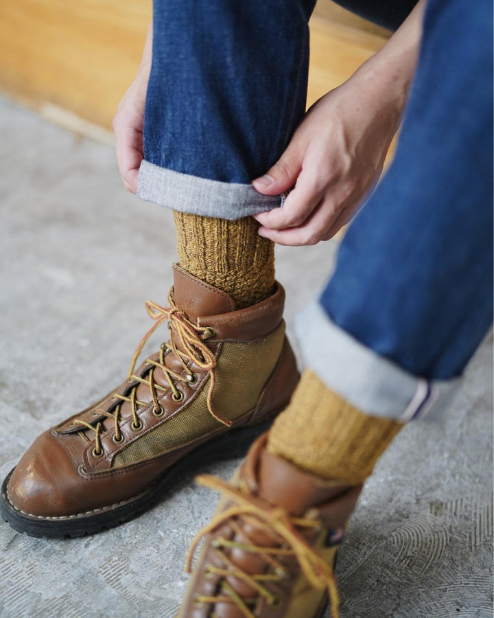 man wearing brown leather boots and mustard boot socks on a concrete floor.