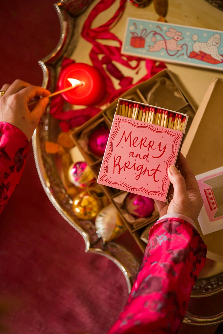 Person lighting a candle with a box of matches labeled 'Merry and Bright' on a decorative table.