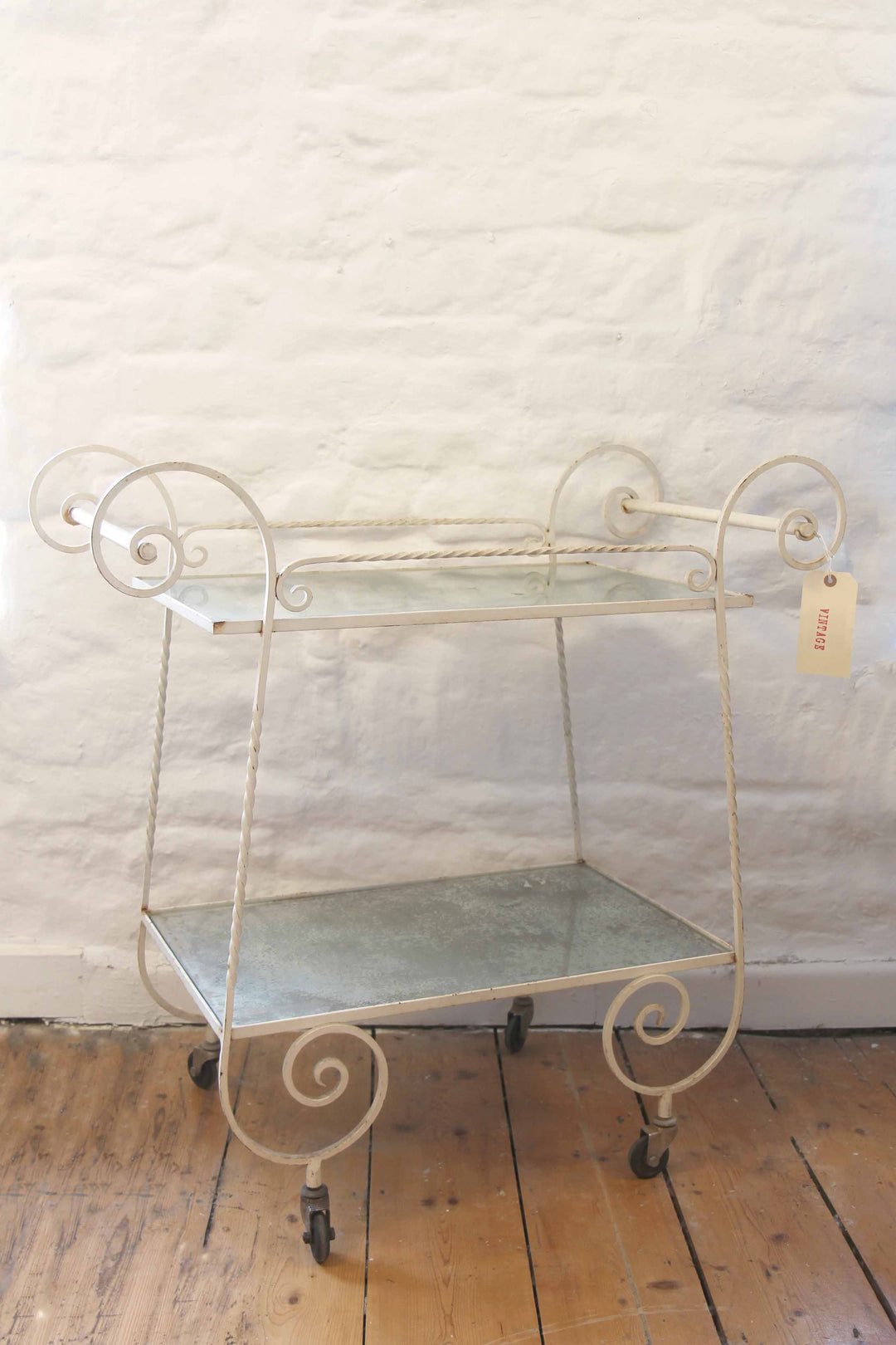Vintage-style metal serving cart with glass shelf against a textured white wall.