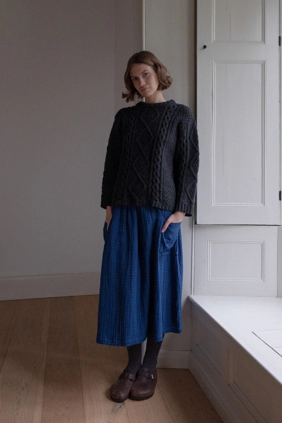 Woman wearing a dark sweater and blue skirt standing in a room with wooden floor and white walls.