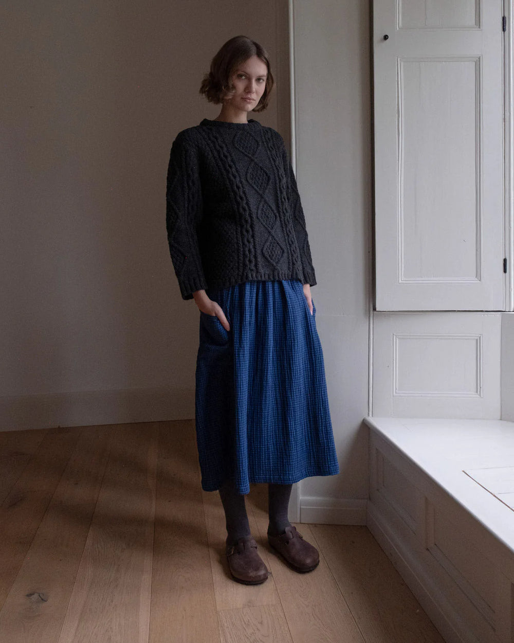 Woman wearing a dark sweater and blue skirt standing in a room with wooden floor and white walls.