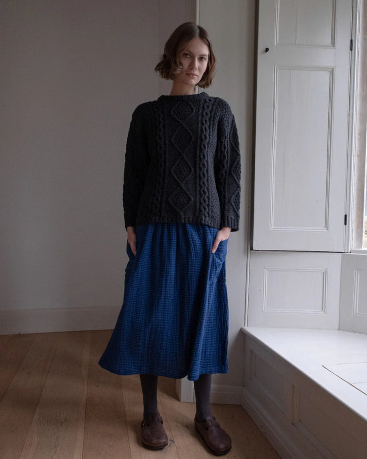 Woman wearing a dark sweater and blue skirt standing in a room with wooden floor and white walls.