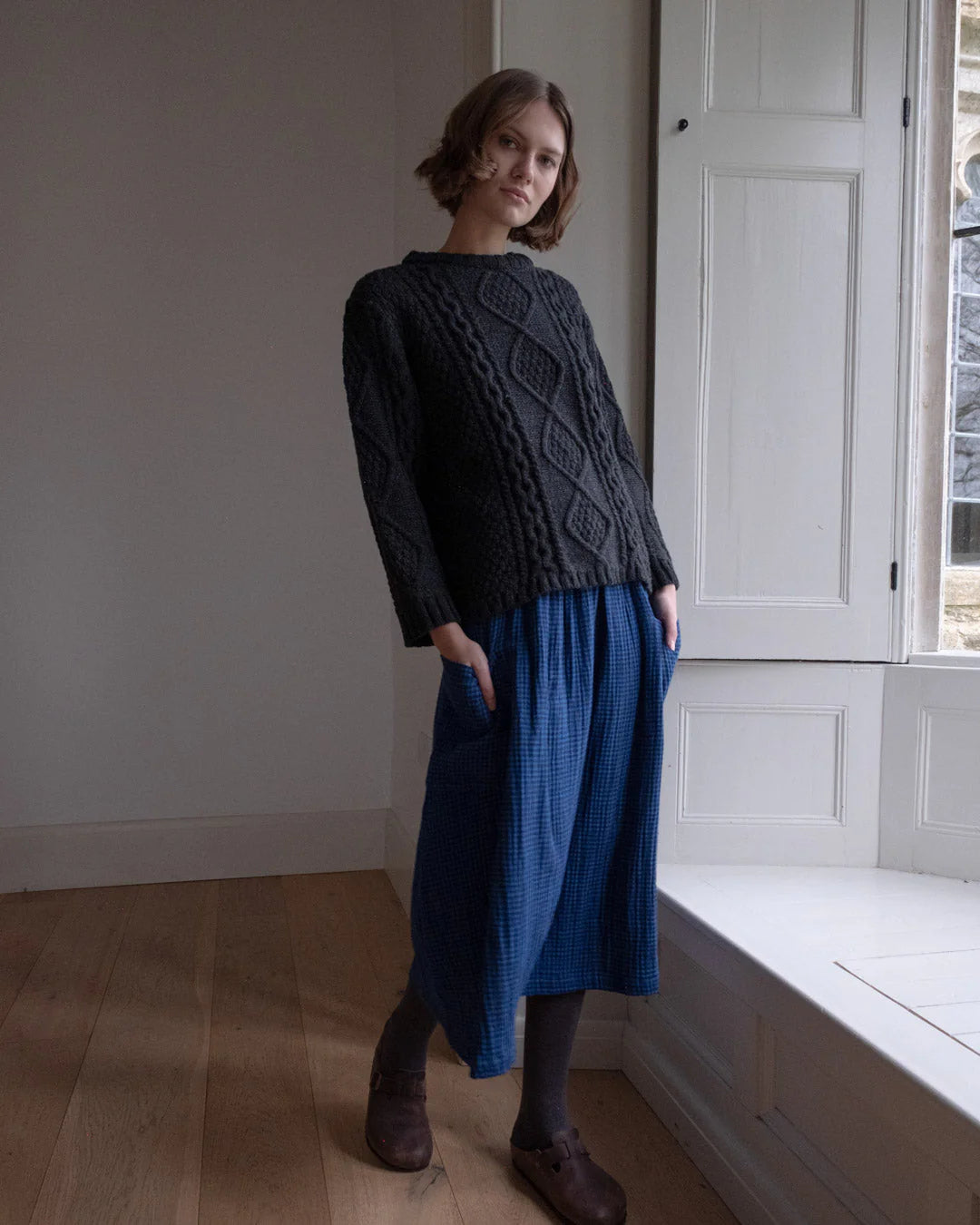 Woman wearing a dark sweater and blue skirt standing in a room with a window.