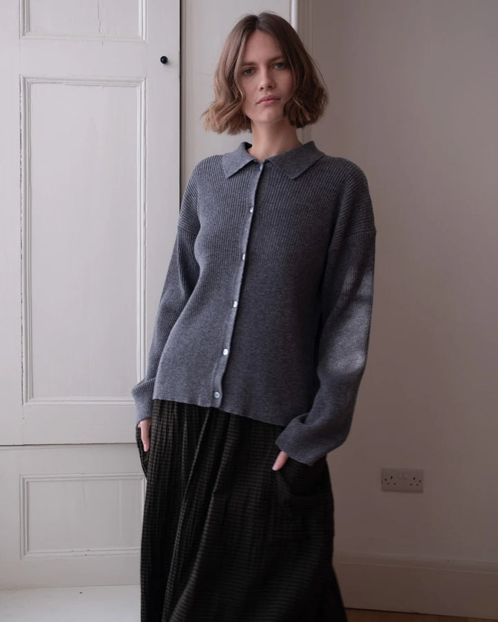 Woman wearing a gray sweater and black pants against a neutral wall.