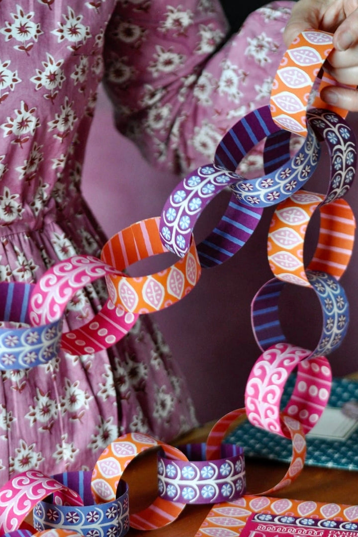 A close-up of a person wearing a pink floral dress holding a long chain of colorful patterned paper rings, likely craft paper or decorative paper. The paper rings feature various designs including stripes, dots, and paisley in orange, pink, purple, blue, and white.